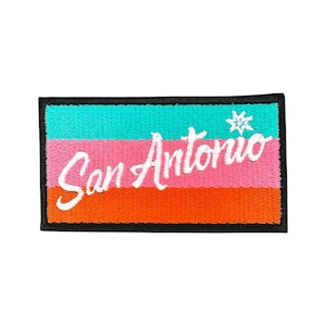 May include: Embroidered patch with the text "San Antonio" in white script. The patch features horizontal stripes in turquoise, pink, and orange, with a black border and a small starburst design.