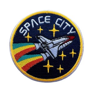 May include: A blue and yellow embroidered patch featuring a space shuttle flying through a rainbow trail. The patch reads "SPACE CITY" in white text.