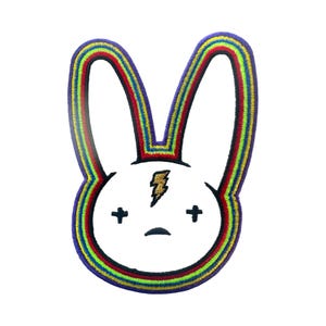 May include: An embroidered patch featuring a white rabbit head with a gold lightning bolt on its forehead, black cross eyes, and a black mouth. The entire shape is outlined with a thick, multi-colored rainbow border of purple, blue, green, yellow, and red.