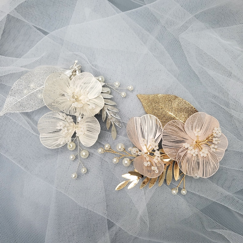 May include: Two floral hair clips with pearl accents. One clip features white flowers with silver leaves and the other features pink flowers with gold leaves.