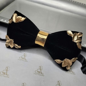 以下が含まれることがあります： 金色の葉のアクセントが付いた黒いベルベットの蝶ネクタイ。蝶ネクタイには、金色の金属製の調節可能なストラップが付いています。