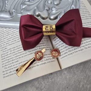 May include: A burgundy bow tie with a gold band featuring Chinese characters, a matching tie clip, and cufflinks. The tie clip and cufflinks have a red circular design with gold accents. The items are displayed on an open book.