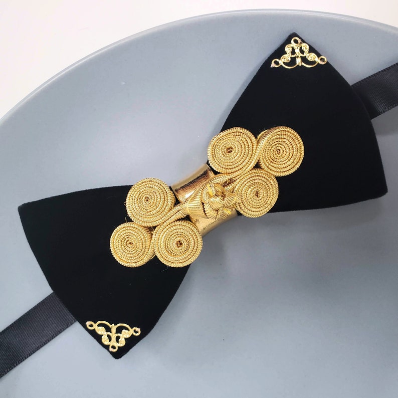 May include: A black velvet bow tie with a gold metal clasp featuring a decorative knot design.