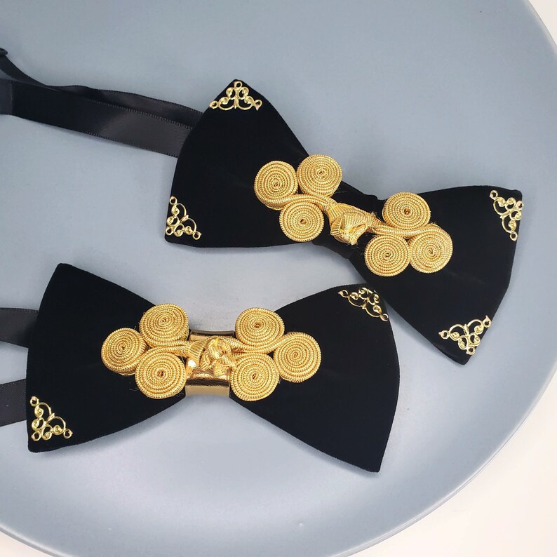 May include: Two black velvet bow ties with gold decorative accents. The bow ties have a unique design with multiple gold circles.