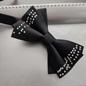 Może przedstawiać: Czarny krawat muszka z srebrnymi kryształkami. Muszka wykonana jest z satyny i ma klasyczny kształt.