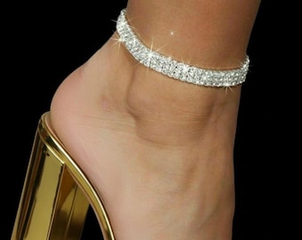 May include: A sparkling, double-layered rhinestone anklet adorns an ankle, paired with a gold, block-heeled sandal. The anklet's shimmering stones catch the light, creating a glamorous effect. The sandal's heel is approximately 3 inches high.