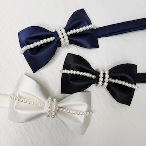 以下が含まれることがあります： ネイビー、ブラック、アイボリーのサテン蝶ネクタイ3つ。各蝶ネクタイは、中央の結び目の周りに模造真珠のストランドで飾られています。蝶ネクタイは、白くテクスチャ加工された表面に表示されています。