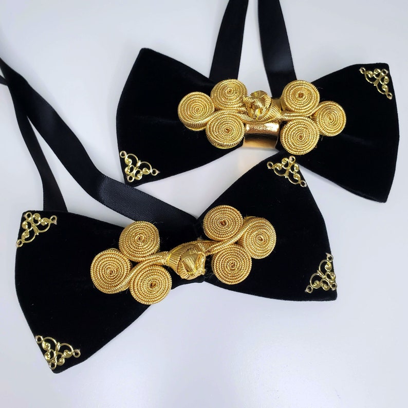 May include: Two black velvet bow ties with gold decorative knots. The bow ties have a satin ribbon for tying around the neck.