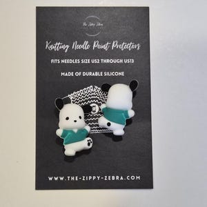 Può includere: Due protezioni per punte di ferri da maglia bianche e verde acqua a forma di personaggi dei cartoni animati, presentate su una carta nera. La carta recita "Knitting Needle Point Protectors" e "Fits Needles Size US2 Through US13."