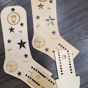 May include: Two wooden sock knitting looms with a zebra design and stars. The looms are made of light brown wood and have a hole for hanging. The text "Zappy Zebra" is engraved on each loom.