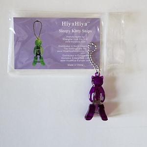 May include: Purple plastic knitting needle tips shaped like a cat with a silver chain. The packaging says "HiyaHiya Sleepy Kitty Snips".