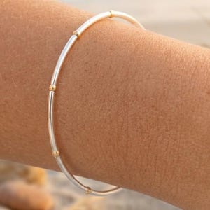 Puede incluir: Una pulsera de plata con detalles dorados. La pulsera, delgada y circular, presenta pequeñas cuentas doradas a intervalos regulares. La pulsera se muestra en el brazo de una persona.