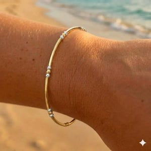 May include: A gold bracelet with silver accents is worn on a wrist. The bracelet has a thin, curved design with small silver beads and a gold band. The background shows a beach with the ocean and sky.