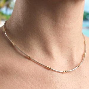 Puede incluir: Un delicado collar con una fina cadena plateada con cuentas doradas espaciadas uniformemente. El collar se lleva alrededor del cuello, mostrando un diseño minimalista. Las cuentas añaden un toque de elegancia a esta joya sencilla pero elegante.