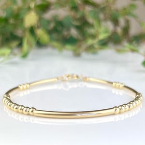 May include: A delicate gold bracelet featuring small, round beads and a smooth, straight bar. The bracelet has a simple clasp. It is displayed on a white surface, with a blurred green background.