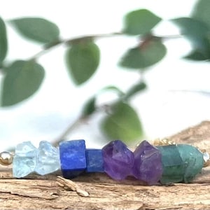 May include: A gold chain necklace with seven gemstone beads in a rainbow color gradient. The beads are aquamarine, lapis lazuli, amethyst, amethyst, and green aventurine.
