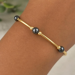 May include: A gold-toned bangle bracelet featuring three dark gray spherical beads spaced evenly along the thin metal band. The design incorporates small gold-toned spacers on either side of each bead, creating a simple and balanced aesthetic.