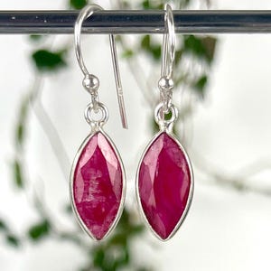 May include: Silver drop earrings featuring marquise-cut, deep red gemstones. The gemstones are set in a silver bezel, and the earrings have a simple hook closure. The earrings are displayed against a blurred background.