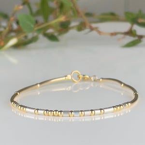 Op de afbeelding: Delicate gouden armband met afwisselend gouden en zilveren kralen. De armband heeft een eenvoudig ontwerp met een sluiting. De sieraden worden gepresenteerd op een wit oppervlak met een vage groene achtergrond.