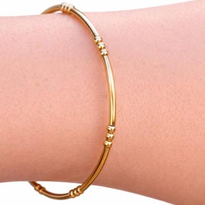 May include: A thin, polished gold-tone bangle bracelet featuring a smooth, circular wire design. The bracelet is accented with small, evenly spaced clusters of three tiny gold-tone beads integrated directly into the band at regular intervals.