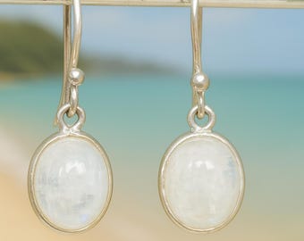 May include: A pair of silver drop earrings with oval, white moonstone cabochons. The earrings have a simple, elegant design with a hook closure. The background is a blurred image of a beach and ocean.