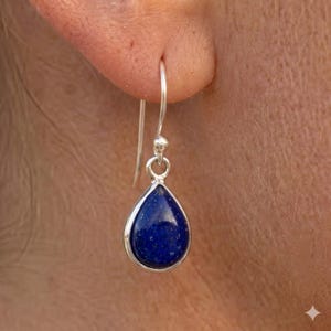 May include: A single silver earring with a teardrop-shaped, deep blue stone. The stone is set in a silver bezel and suspended from a silver hook. The earring is shown close up against a blurred background.