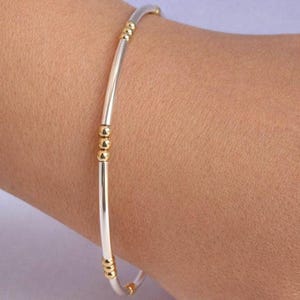 May include: A silver and gold bangle bracelet. The bracelet has a silver band with gold detailing. The gold detailing consists of small, round beads. The bracelet is worn on a wrist.