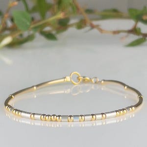 May include: A delicate gold and silver beaded bracelet. The bracelet features small, round gold beads and rectangular silver beads, connected by a thin gold chain. The clasp is a simple gold hook and eye closure. The bracelet is designed for a minimalist aesthetic.
