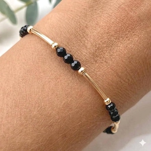 May include: A beaded bracelet featuring alternating segments of small, faceted black beads and smooth, cylindrical gold-toned metal bars. The design consists of repeating patterns of three black beads separated by single gold-toned metal tubes.