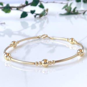 Può includere: Un braccialetto dorato dal design delicato. Il braccialetto presenta diverse perle rotonde e lucide di varie dimensioni infilate lungo una sottile fascia curva. Il braccialetto è esposto su una superficie bianca con uno sfondo verde sfocato.