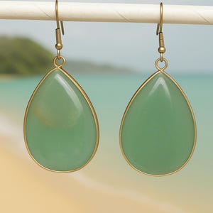 May include: Teardrop earrings featuring pale green stones with gold-coloured metal edging. The earrings are suspended from gold-coloured hooks. The background is a blurred beach scene.