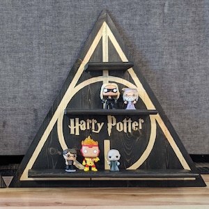 May include: A black triangular shelf with three shelves, featuring a gold and black Harry Potter symbol. The shelf is decorated with five small figurines, including Harry Potter, Hermione Granger, Ron Weasley, and two other characters.
