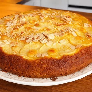 Op de afbeelding: Een ronde appeltaart met een goudbruine korst, belegd met dun gesneden appelschijfjes en geschaafde amandelen, geserveerd op een wit bord met een delicaat bloemmotief langs de rand, met een gebakken, getextureerd oppervlak.