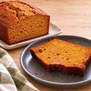 May include: A golden-brown loaf of baked bread on a rectangular white plate, accompanied by a single slice of the same bread served on a round grey plate. A green and white checkered cloth is positioned to the side of the plates.