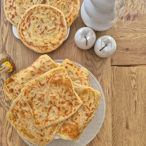 Peut inclure: Un assortiment de pains plats dorés, comprenant plusieurs pains ronds feuilletés de type paratha sur une assiette blanche et une pile de pains carrés pliés de type msemmen sur un tapis tissé rond, présentant une texture grillée et feuilletée.