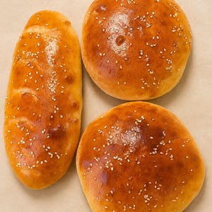 Peut inclure: Trois petits pains dorés à la finition brillante, parsemés de petites graines de sésame blanches. Un pain est de forme allongée et ovale, tandis que les deux autres sont ronds. Le pain présente une croûte lisse et une couleur dorée.
