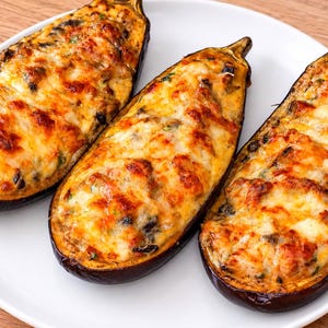 May include: Three halves of roasted aubergine arranged on a white plate, each filled with a savoury mixture topped with melted, golden-brown cheese. The dark purple skins of the aubergines contrast with the bubbly, browned surface of the filling.