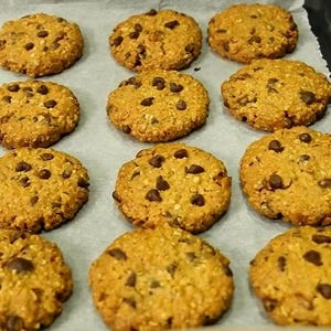 Peut inclure: Un lot de biscuits ronds à l'avoine, dorés, parsemés de pépites de chocolat noir, disposés en rangées sur du papier sulfurisé. Chaque biscuit présente une surface texturée avec des flocons d'avoine visibles et des éclats de chocolat.
