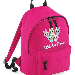Może przedstawiać: Jasnoróżowy plecak z czarnymi paskami i uchwytem. Posiada przednią kieszeń i komorę główną, obie z czarnymi zamkami błyskawicznymi. Projekt zawiera kreskówkowe króliki i napis "Lilah-Rose" w ozdobnej czcionce.