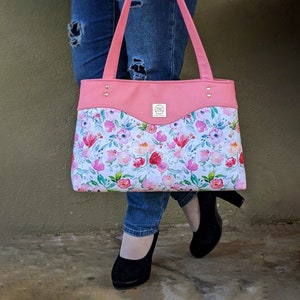 Op de afbeelding: Een roze tote bag met een bloemenprint en een roze flap met een gouden logo. De tas heeft een witte achtergrond met roze, rode en gele bloemen.