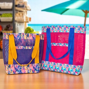 以下が含まれることがあります： 2つのビーチトートバッグ。1つは黄色のハンドルが付いた紫、もう1つは青いハンドルが付いたピンクです。どちらもメッシュデザインとカラフルなパターンです。背景にはパラソルのあるビーチシーンが写っています。