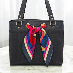 Pode incluir: Bolsa tote preta com textura tecida na metade inferior e parte superior preta lisa. A bolsa tem duas alças e é adornada com um lenço colorido amarrado em um laço. O lenço apresenta listras vermelhas, azuis, amarelas e rosas.