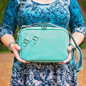 May include: A turquoise crossbody bag with silver-tone hardware. The bag features a rectangular shape with a zipper closure and a patterned strap. The bag is held by a person wearing a blue patterned dress.