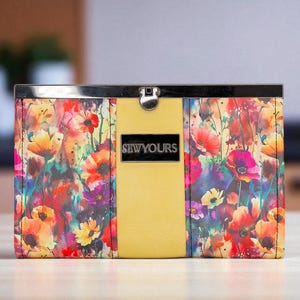 Op de afbeelding: Een rechthoekige bloemenportemonnee met een metalen sluiting. De portemonnee heeft een levendig aquarelontwerp van rode, gele en paarse bloemen. Een geel paneel is gecentreerd op de voorkant, met de tekst "SEWYOURS".