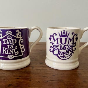 May include: Two ceramic mugs with curved handles. One mug is purple with the text "DAD is KING" in white. The other mug is white with purple text that reads "MUM is Queen". Both mugs have a cream-colored base.