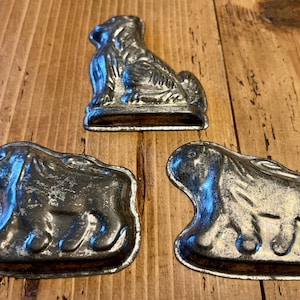 May include: Three antique metal molds in the shape of animals. One mold is a dog, and the other two are rabbits. The molds are silver in color and sit on a wooden surface.