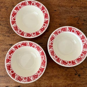 May include: Three white ceramic bowls with a red floral pattern around the rim. The bowls are arranged on a wooden surface. The floral design is a repeating pattern of stylized flowers and leaves.