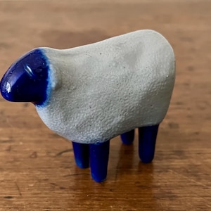 May include: A small ceramic sheep figurine featuring a textured, matte light gray body and a smooth, glossy deep blue head and legs. The sheep stands on four legs and is depicted in a side profile view, showcasing the contrast between the rough gray surface and the shiny blue glaze.