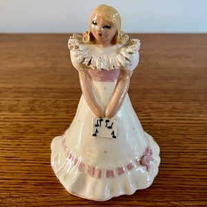 May include: A ceramic figurine of a girl wearing a white dress with pink accents and a white lace collar. She is holding a sheet of music with musical notes printed on it.