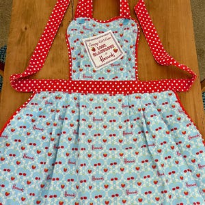May include: A red and white polka dot apron with a blue and white patterned skirt. The skirt has a repeating pattern of strawberries and the Harrods logo. The apron has a small white tag that says "Every Girl Can Look Glamorous at Harrods".